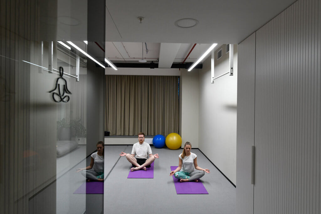 "A diverse group of individuals, dressed in comfortable workout attire, are stretching and finding their balance while practicing yoga on soft purple mats in a serene lounge area of an office setting. The surrounding is decorated with calming and natural elements, a wooden floor and plants in the background, indicating a dedicated space for mental and physical well-being.