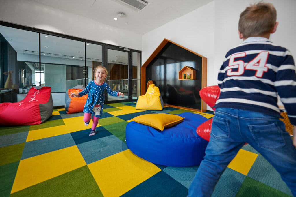 "A group of children, of different ages, are having fun and play in a spacious 6x6 meter room, decorated with lively colors. The floor is covered with a colorful carpet, with different shapes and hues. The children are seen playing with different toys and games, from building blocks to dolls. The room is filled with laughter and smiles, which indicate that this is a happy and fun place for kids to spend their time. The image portrays a dedicated and stimulating space for children to learn and grow, as well as foster creativity and imagination.